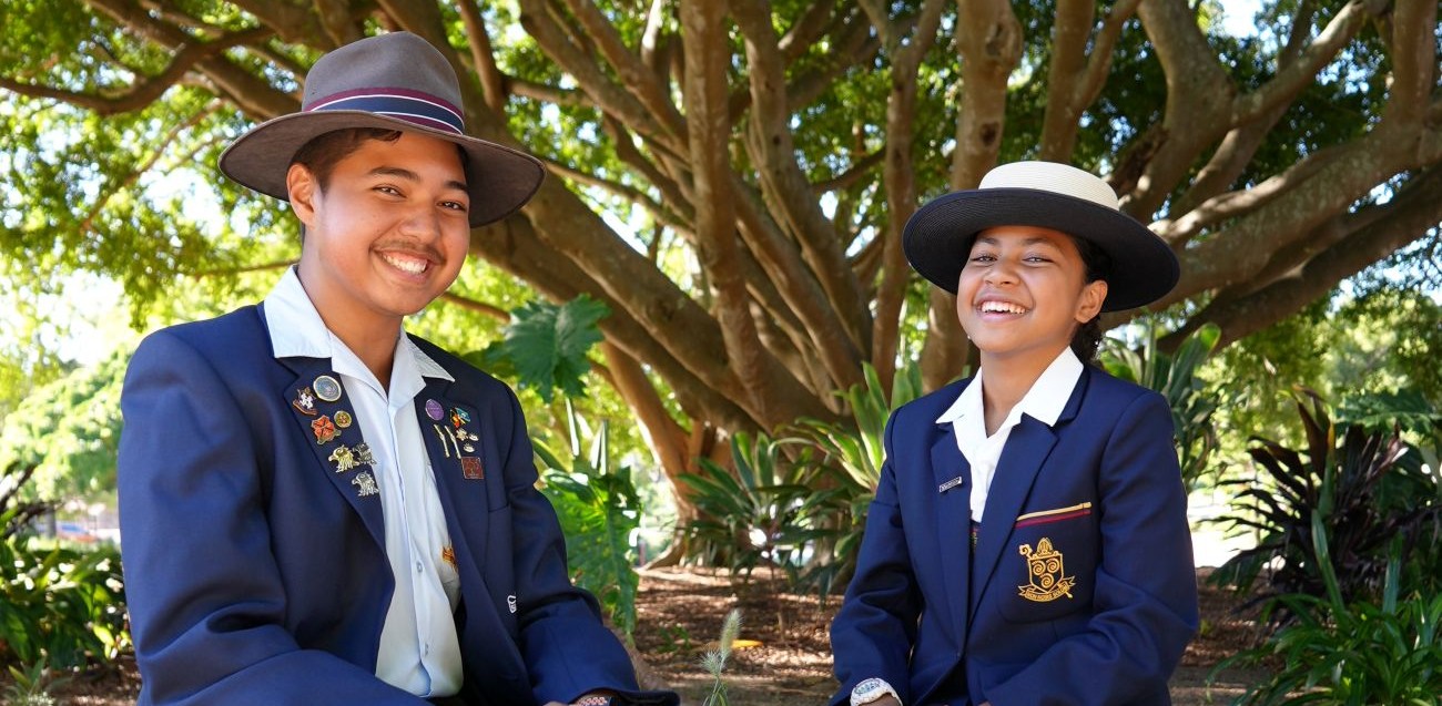 Two students in navy blazers with badges sit outdoors under a large tree smiling at the camera   yalari   yalari