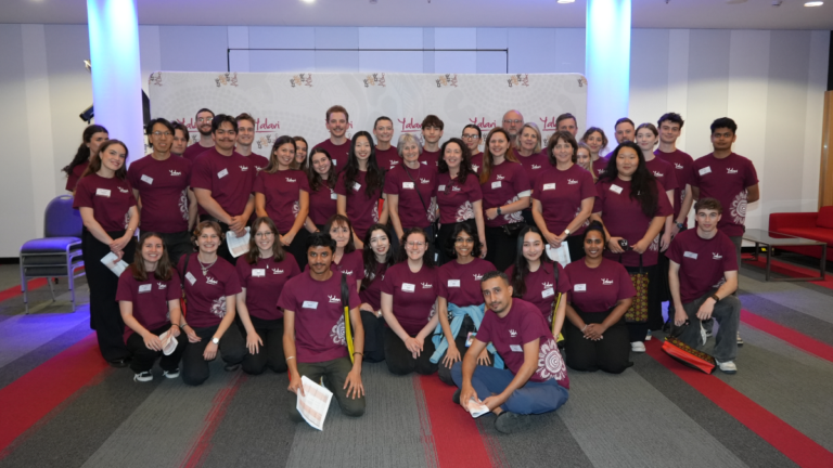 Group of around 40 people in maroon event shirts posing for a photo in a conference space with a branded backdrop and name tags on their chests   yalari   yalari