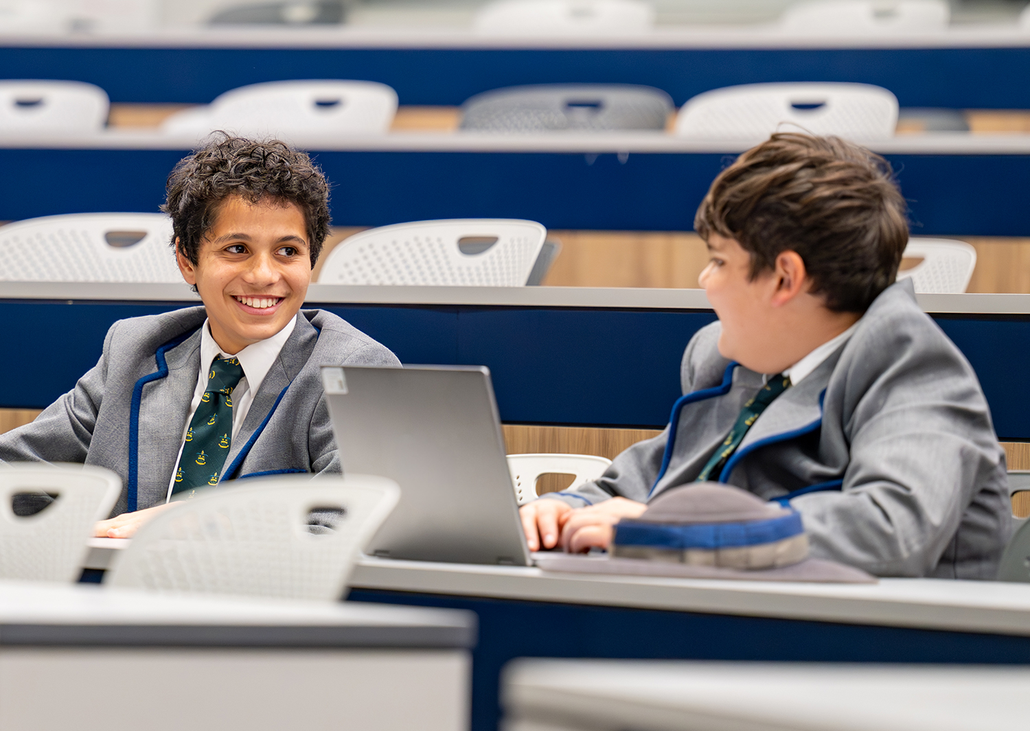 Two schoolboys in gray blazers and green patterned ties smile at each other while seated in a lecture hall with a laptop open on the desk in front of them   yalari   yalari