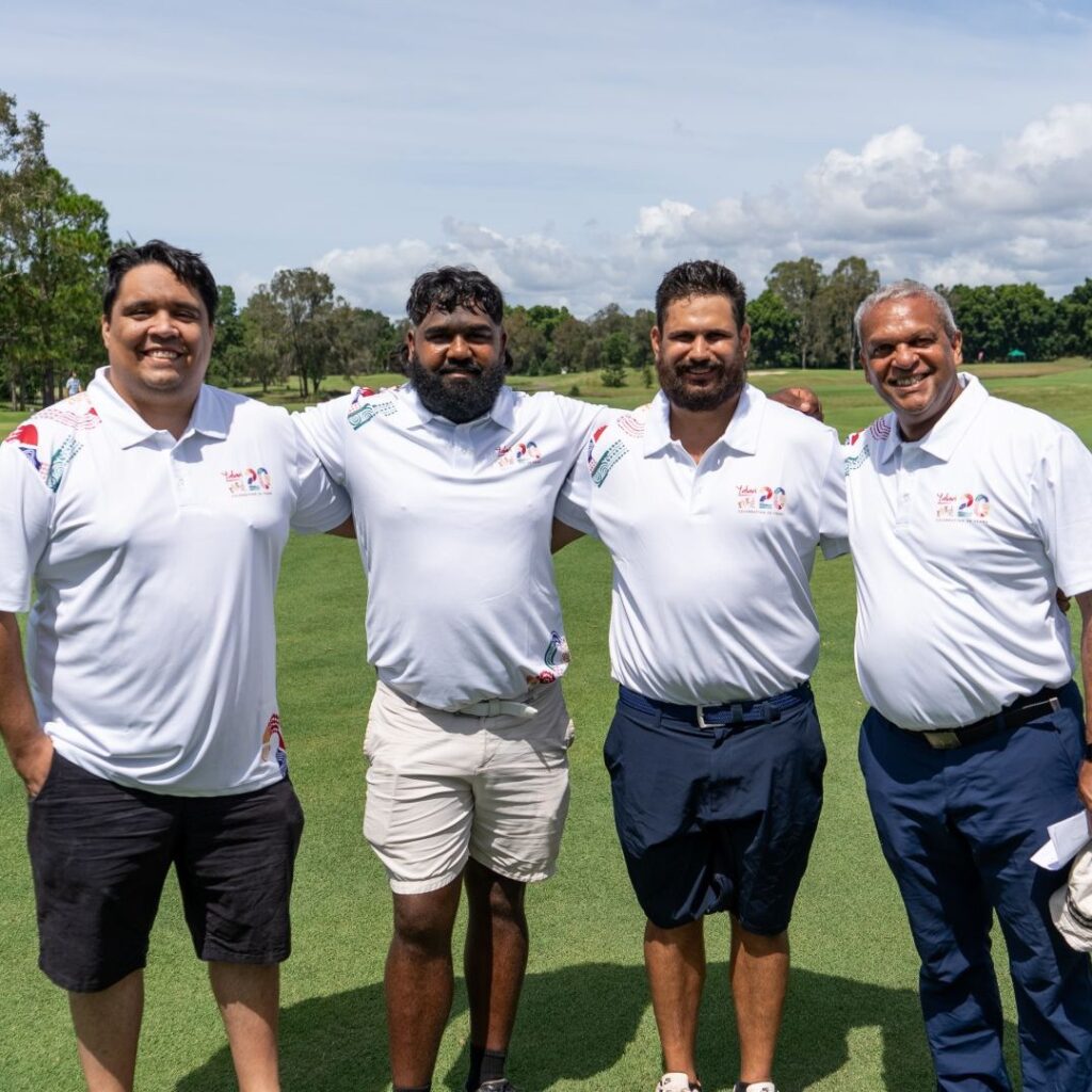Four men in white polo shirts stand with arms around each other on a sunny golf course.
