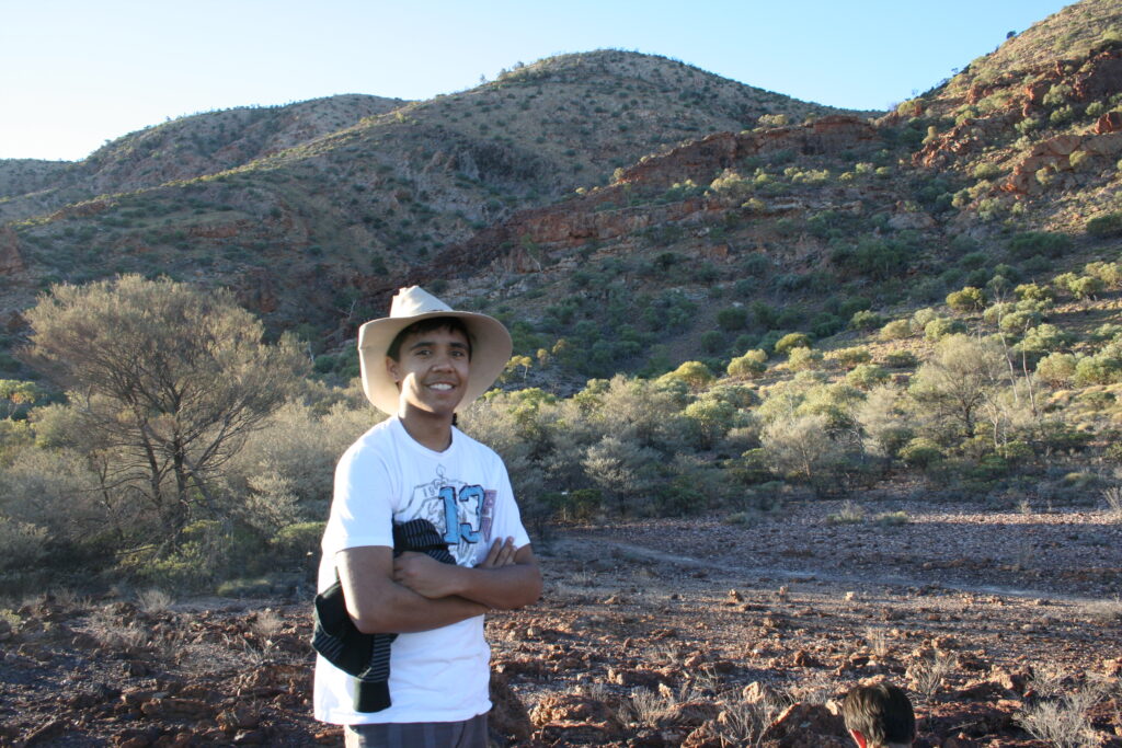 Will munro 2009 - Yalari Man wearing a wide beige hat and white shirt stands with arms crossed in a rocky desert landscape hills in the background yalari