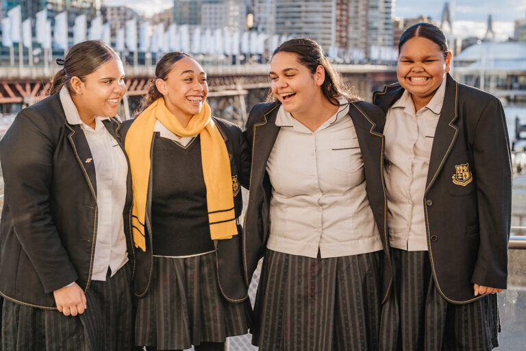 Four high school girls in uniforms stand with arms around each other and smile on a waterfront boardwalk   yalari   yalari