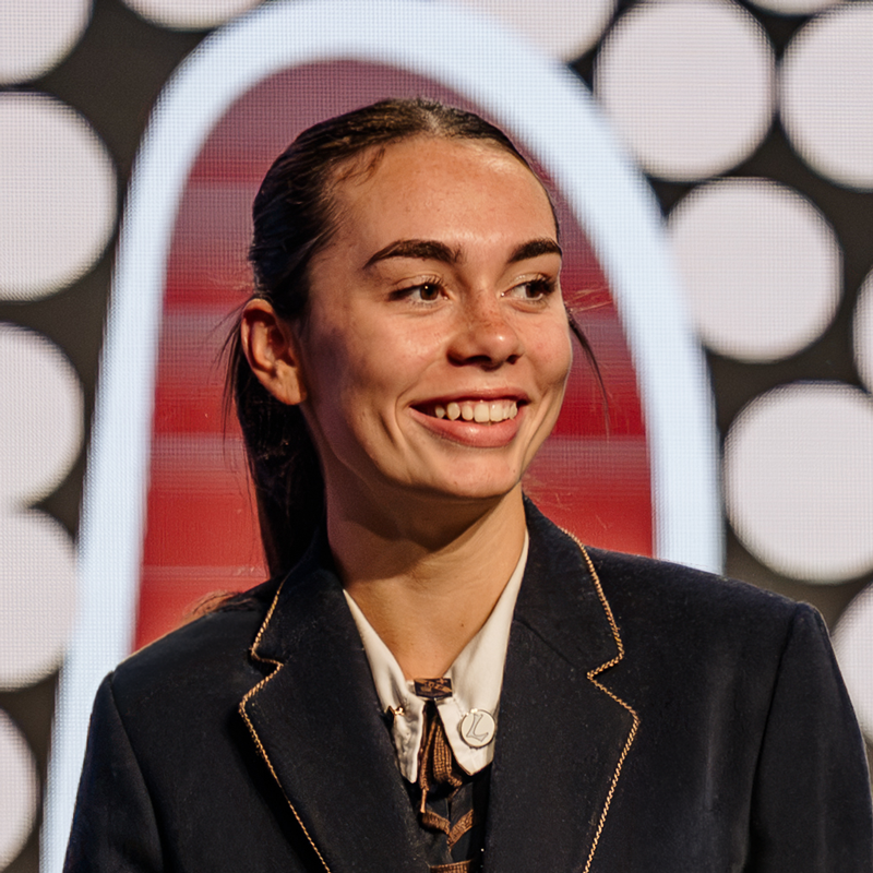 Smiling woman in a dark blazer with light shirt on a stage a rounded light backdrop behind her   yalari   yalari