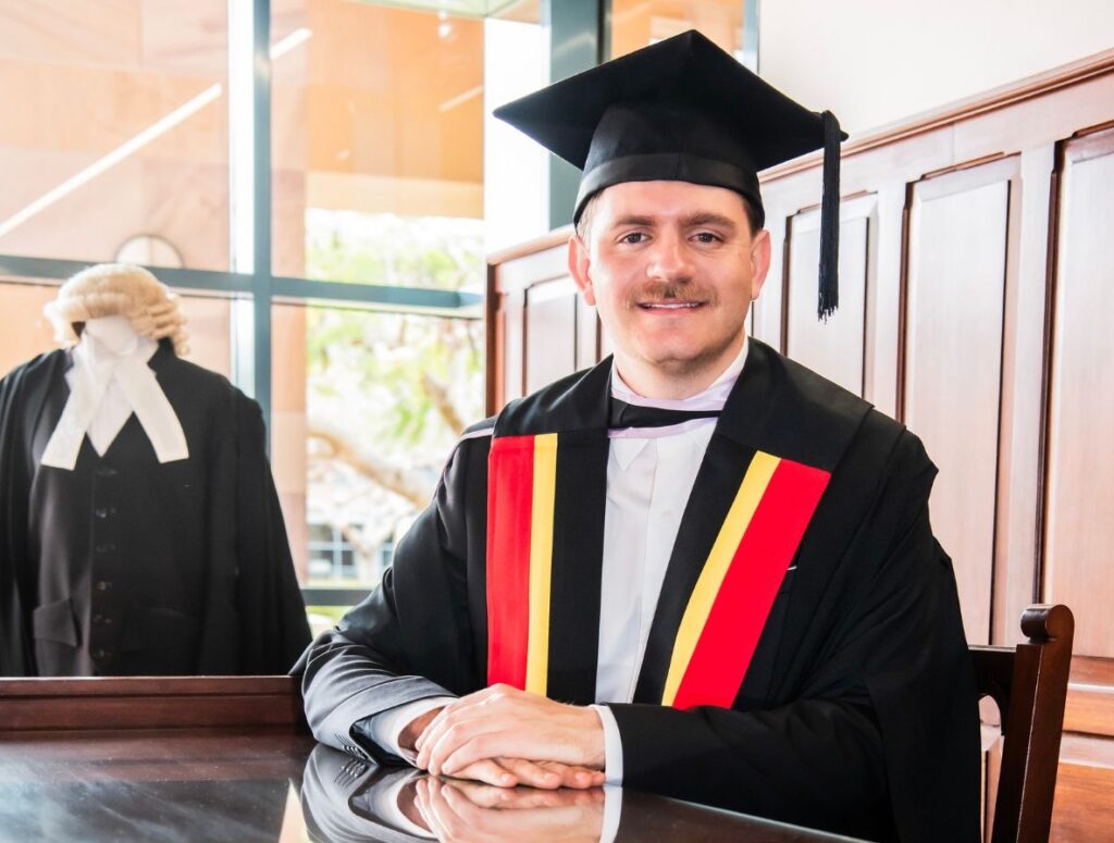 Graduate in mortarboard and black gown with a redyellowblack sash sits at a wooden desk a judges wig stands in the background   yalari