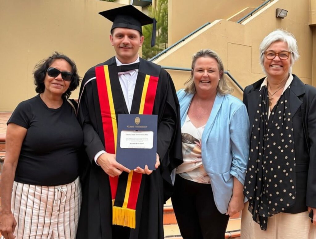 Graduate in cap and gown holding a diploma flanked by three smiling adults at an outdoor staircase   yalari