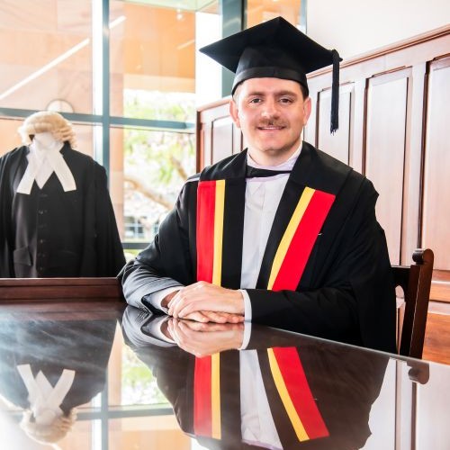 Graduate in cap and gown with a colorful red yellow stole sits at a polished desk a courtroom wig is visible in the background   yalari   yalari