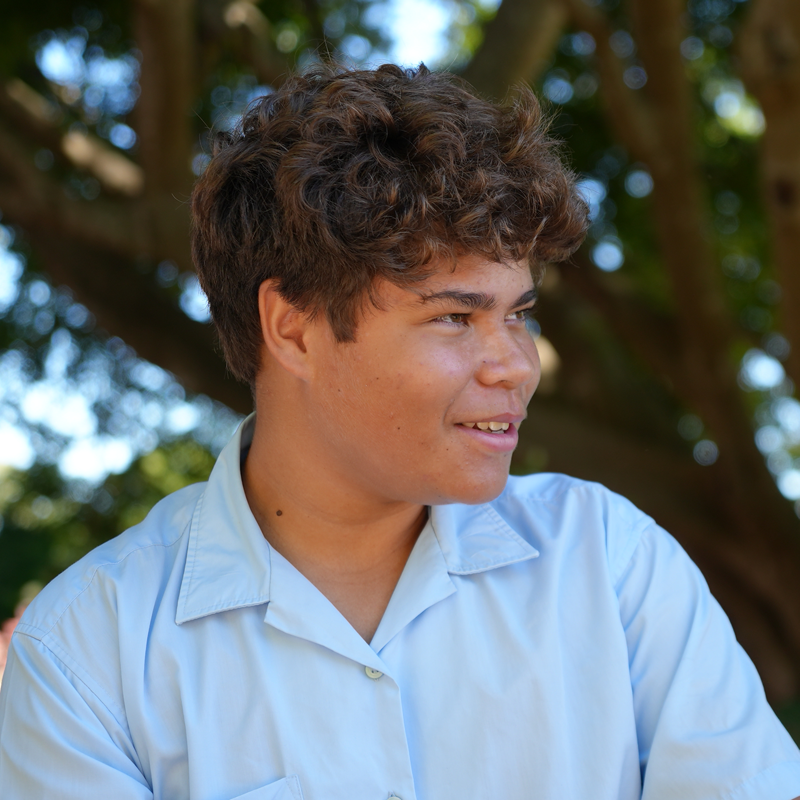 Young person with curly brown hair in a light blue shirt outdoors among trees looking to the right with a gentle smile   yalari   yalari