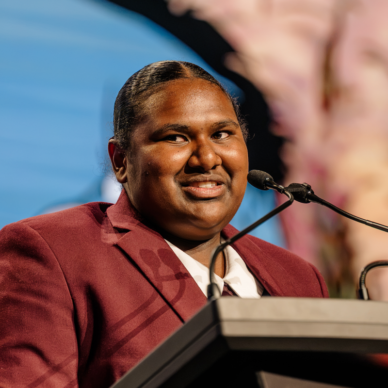 Person in a maroon blazer speaking at a lectern with a microphone against a blue backdrop at a formal event   yalari   yalari