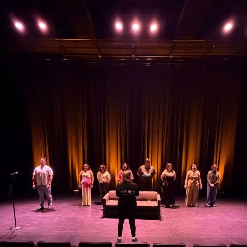 Eight performers stand in a line on a dimly lit stage with warm amber backlights a conductor faces them from the front a sofa centered on stage   yalari   yalari