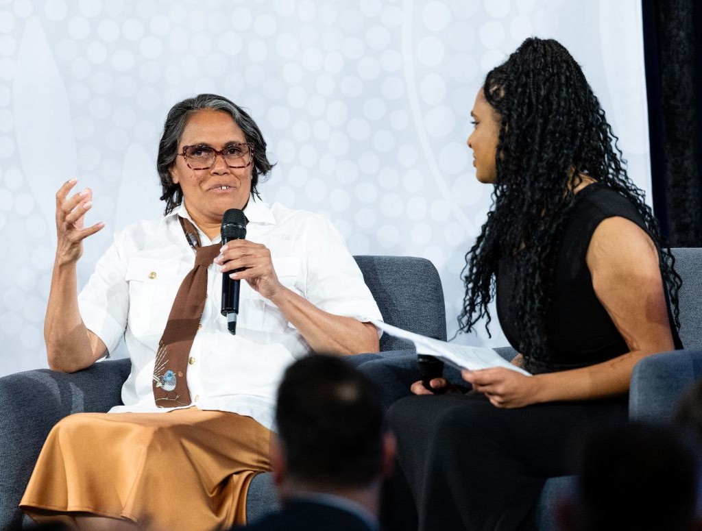 Two women sit on a panel stage one in a white shirt and brown scarf speaks into a handheld mic while the other in black attire holds papers and listens   yalari