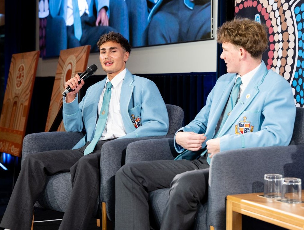 Two students in light blue blazers sit on stage in armchairs during a panel discussion one speaks into a microphone while the other listens   yalari