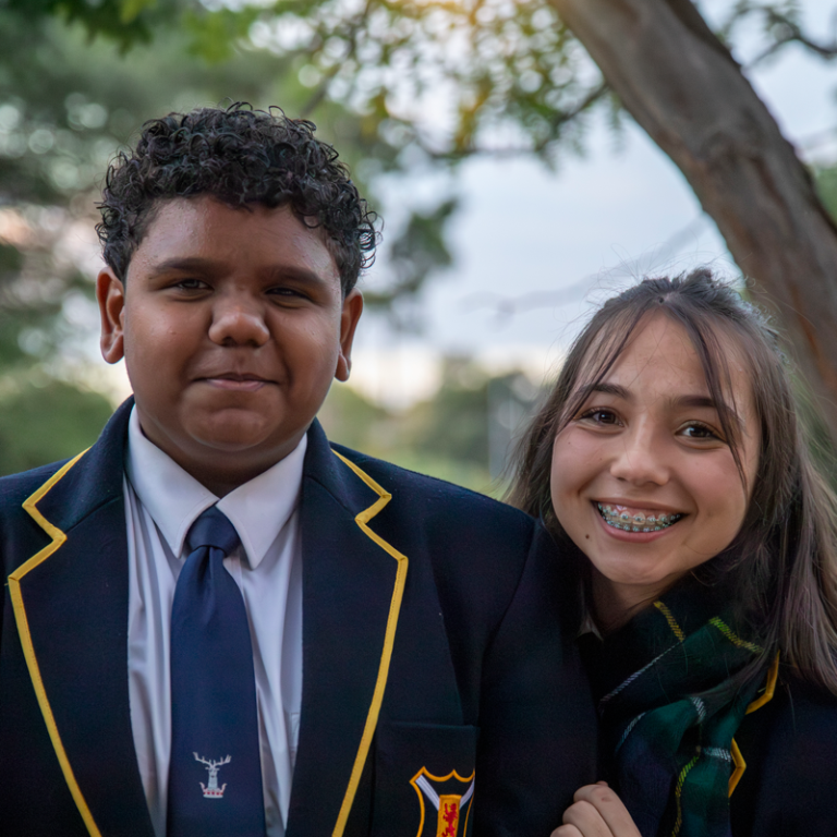 Two students in school uniforms pose together outdoors the boy on the left wearing a navy blazer with yellow trim and tie the girl on the right smiling with braces   yalari   yalari
