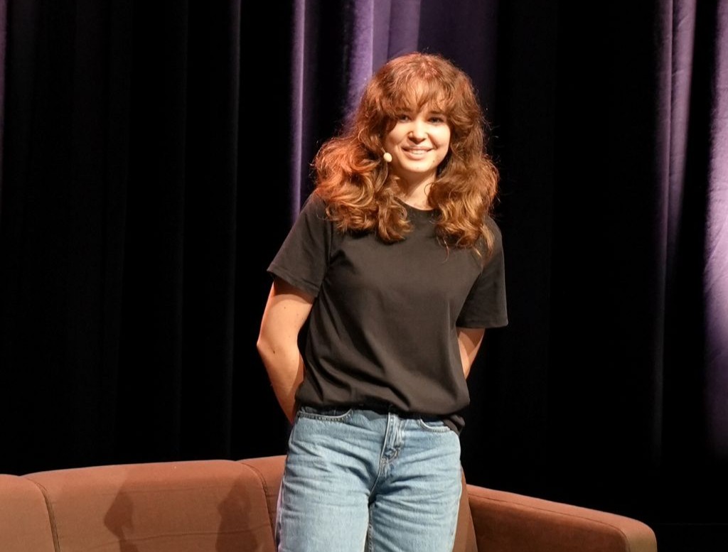 Woman on stage with a headset mic smiling wearing a black t shirt and jeans standing in front of purple curtains and a brown sofa   yalari