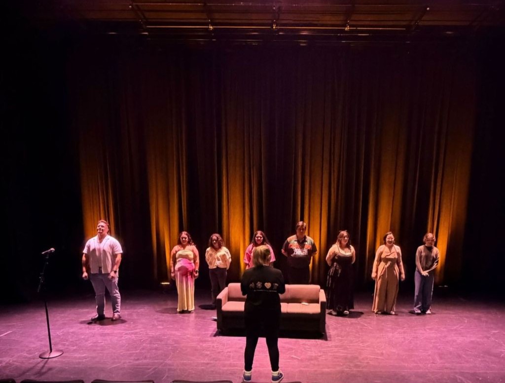 Nine performers stand in a line on a dark theater stage with amber lights facing a conductor in front of a sofa   yalari